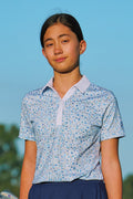 A young woman wearing a light blue and white floral patterned short-sleeve polo shirt with a light blue collar, set against a clear blue sky background.