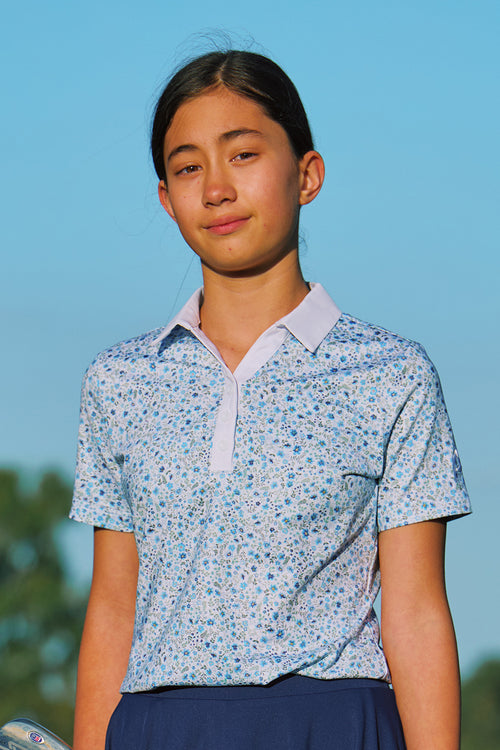 A young woman wearing a light blue and white floral patterned short-sleeve polo shirt with a light blue collar, set against a clear blue sky background.