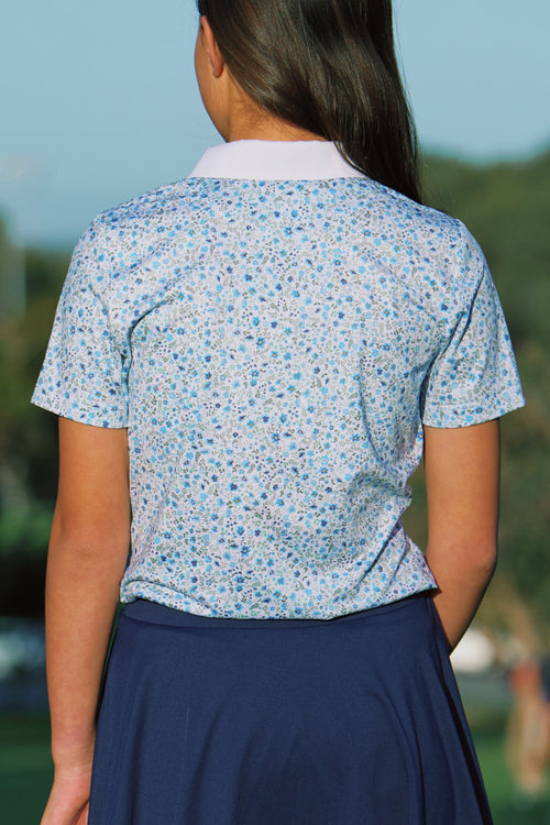 A girl viewed from behind wearing a short-sleeve collared golf polo shirt with a dense small floral print in light blue, medium blue, and white, paired with a dark navy skirt. The background is a blurred outdoor landscape.