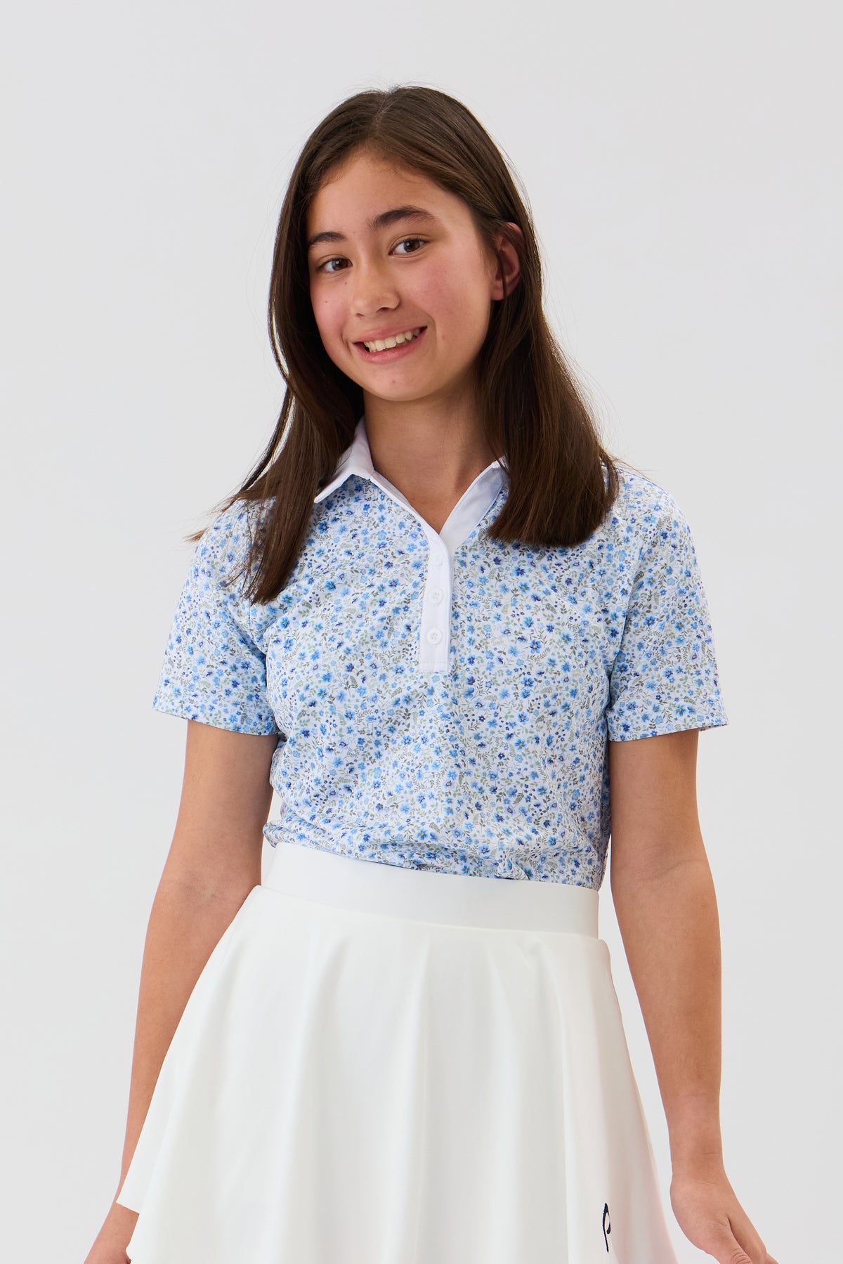 A young girl with long brown hair smiling, wearing a light blue and white floral patterned short-sleeve polo shirt with a white collar and a white skirt, set against a plain light gray studio background.