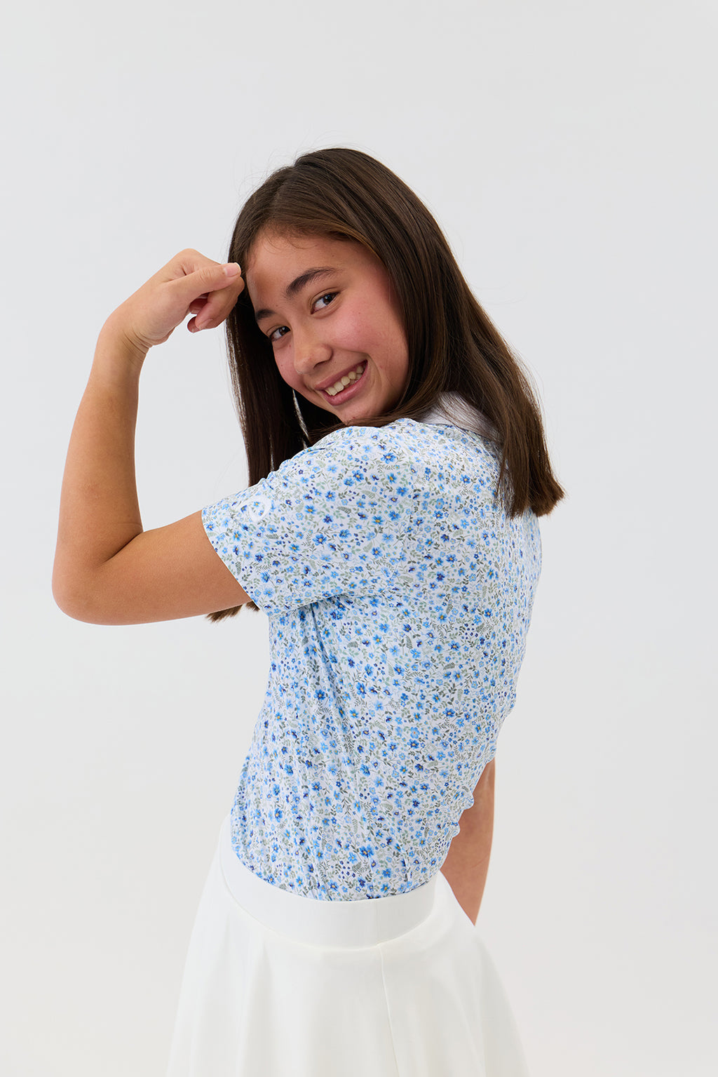 A 11 year old girl  with long brown hair is shown from the back and side, wearing a light blue and white floral print short-sleeve top and a white skirt, posing against a solid white studio background.