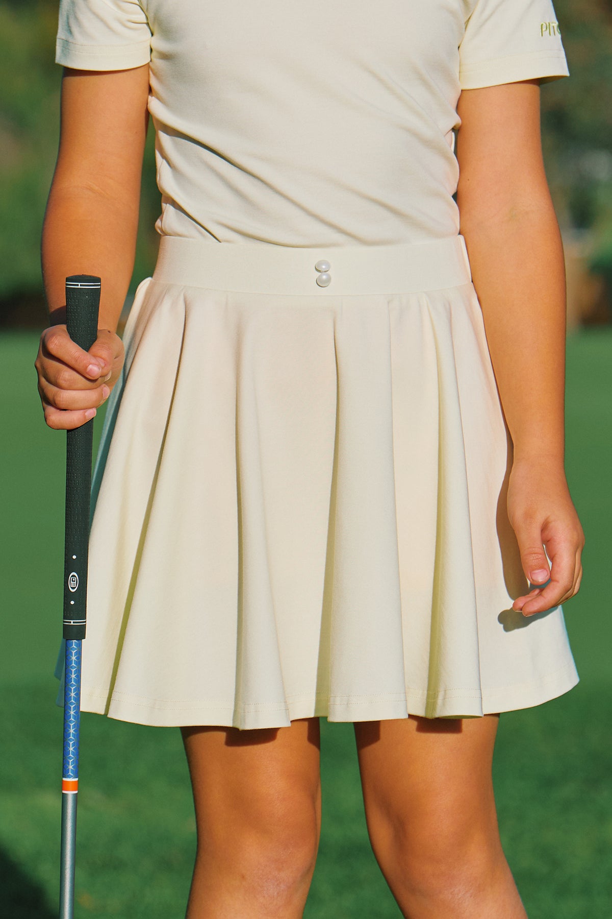 A close-up of a junior girl golfer wearing a cream-yellow colored short-sleeve polo shirt and matching pleated golf skirt, holding a golf club on a green grass field.