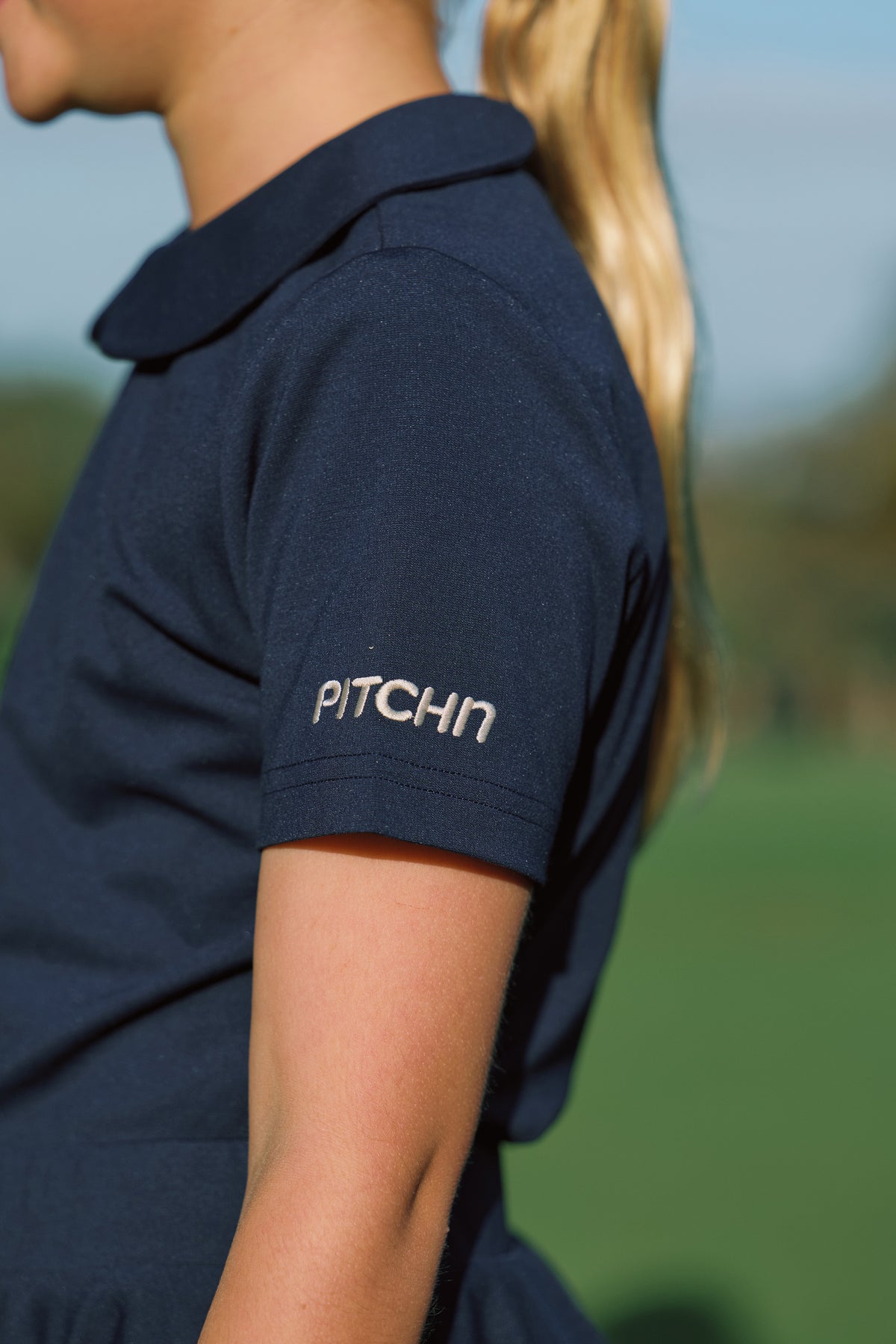 A close-up side profile of a girl wearing a navy blue golf polo shirt with 'PITCHN' embroidered in white on the sleeve, set against a blurred green outdoor background.