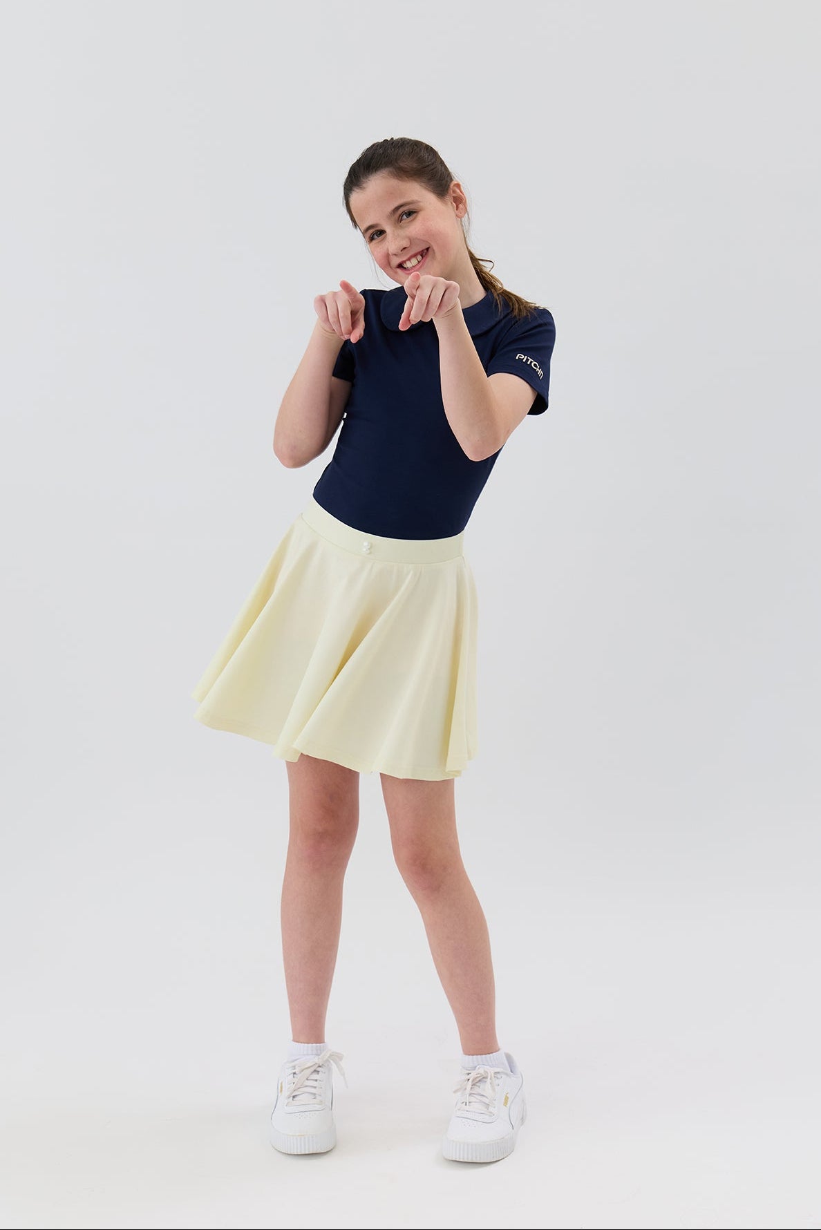 A young girl posing in a navy blue short-sleeve athletic golf top and a light yellow pleated skirt, paired with white sneakers, against a plain white studio background.