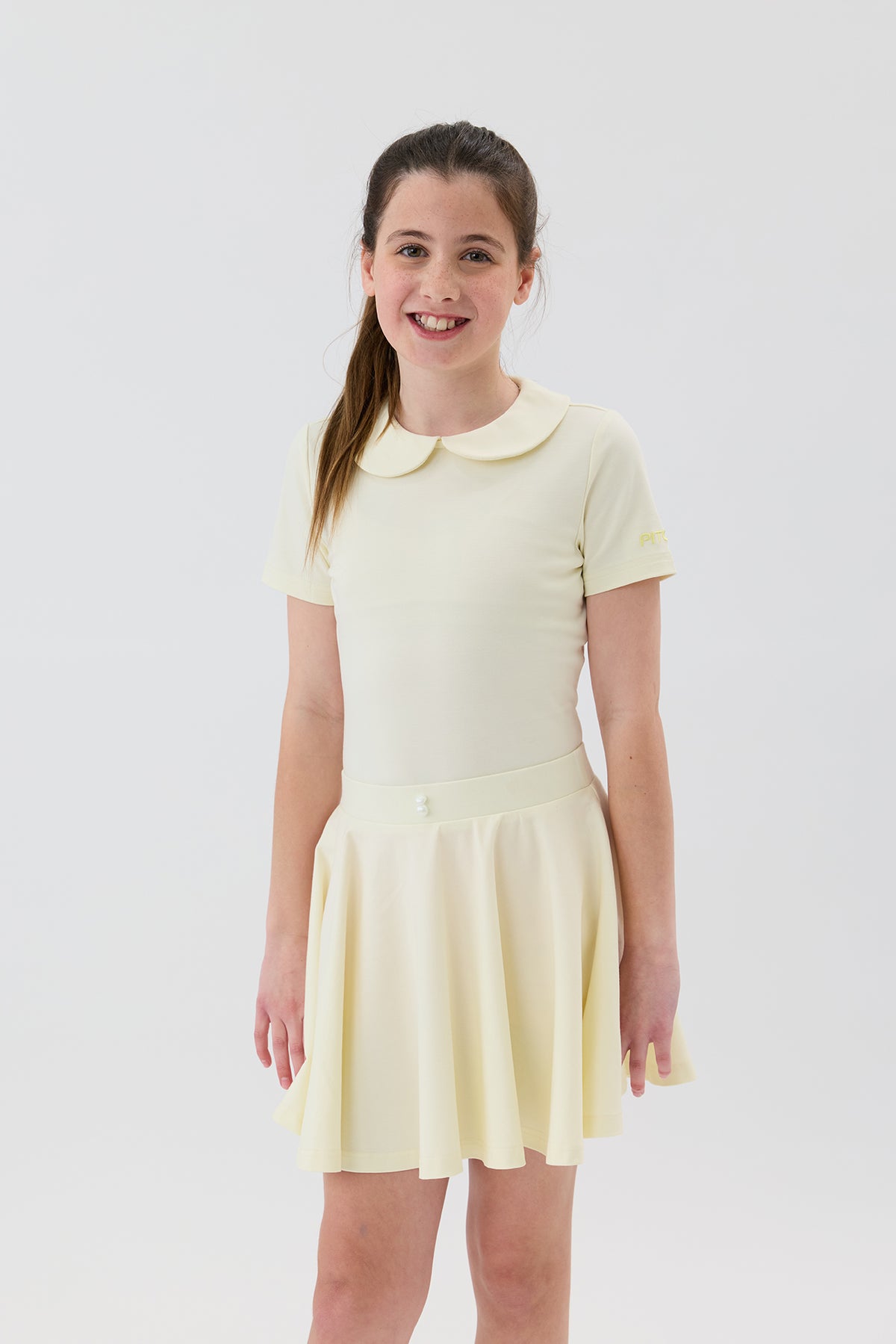 A young girl golfer with her hair in a ponytail stands against a light gray studio background wearing a pale yellow golf top 
