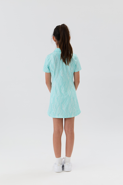 A girl standing with her back to the camera, wearing a light mint green short-sleeve collared golf dress with a subtle tonal leaf pattern, paired with white socks and white sneakers, against a plain white studio background.