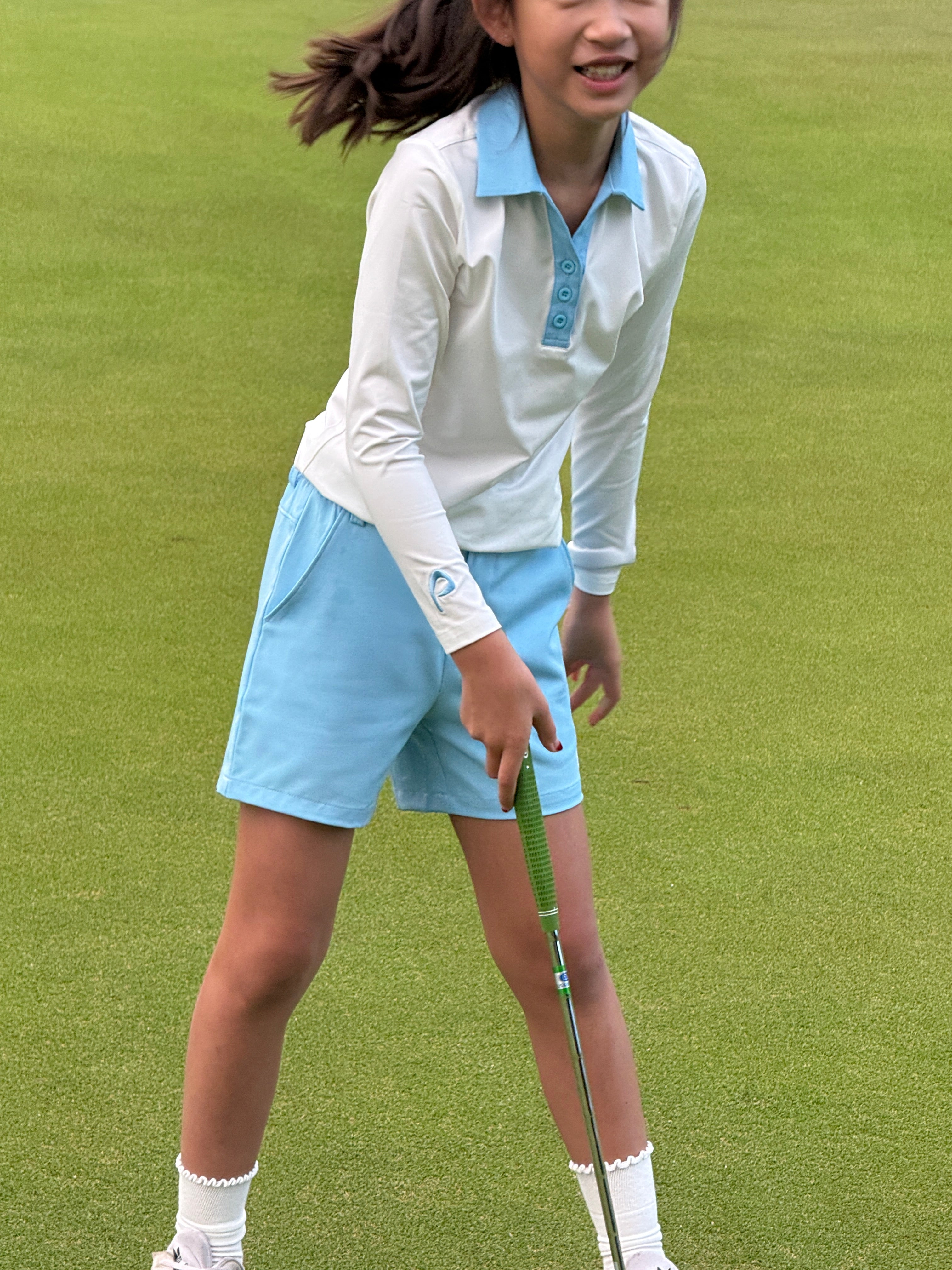 Girl in a white longsleeve polo and blue skirt holding a putter on  golf course