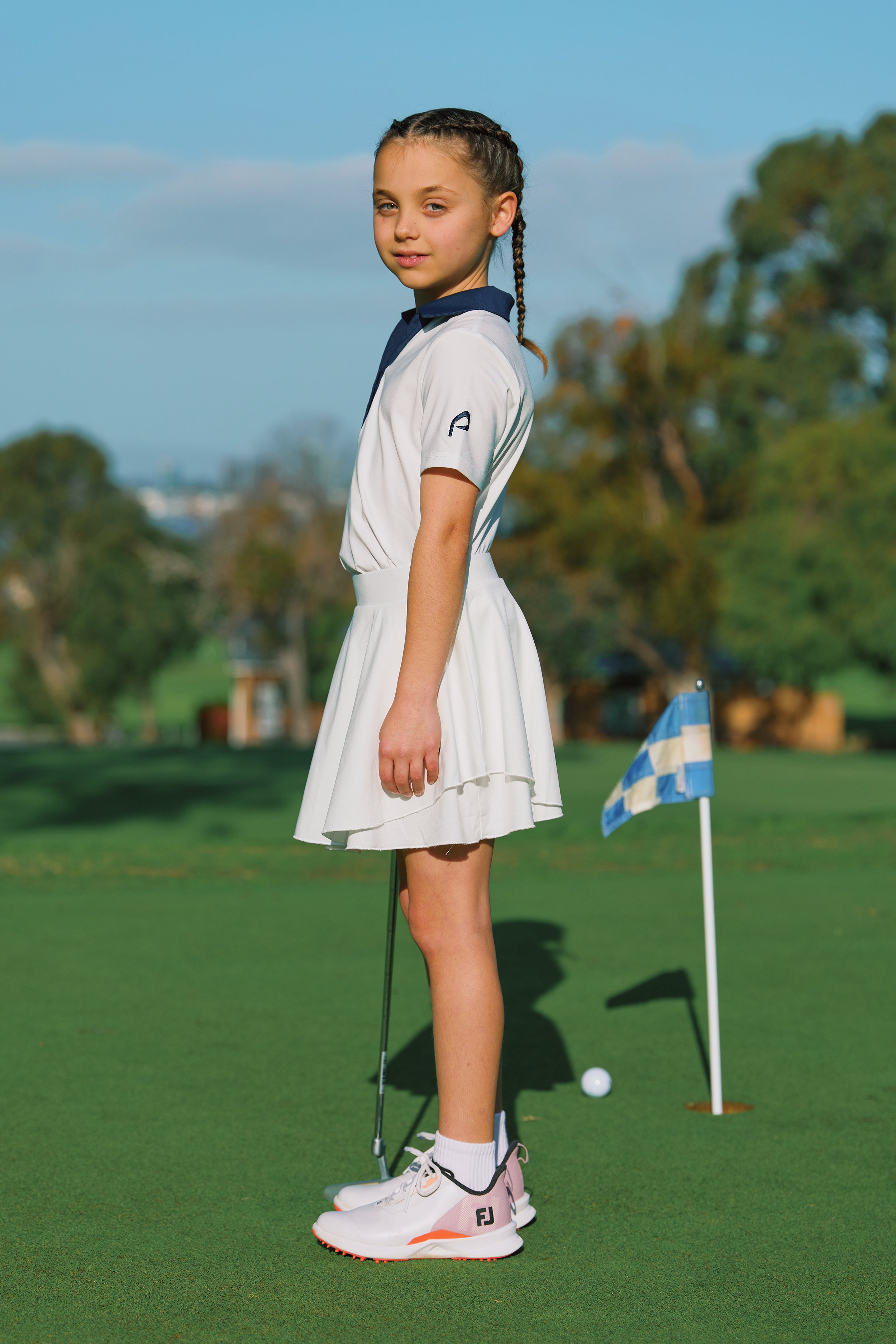 A young girl stands on a golf course wearing a white golf dress with a navy collar, white golf shoes, and holding a putter. A blue and white checkered flag is visible in the background.