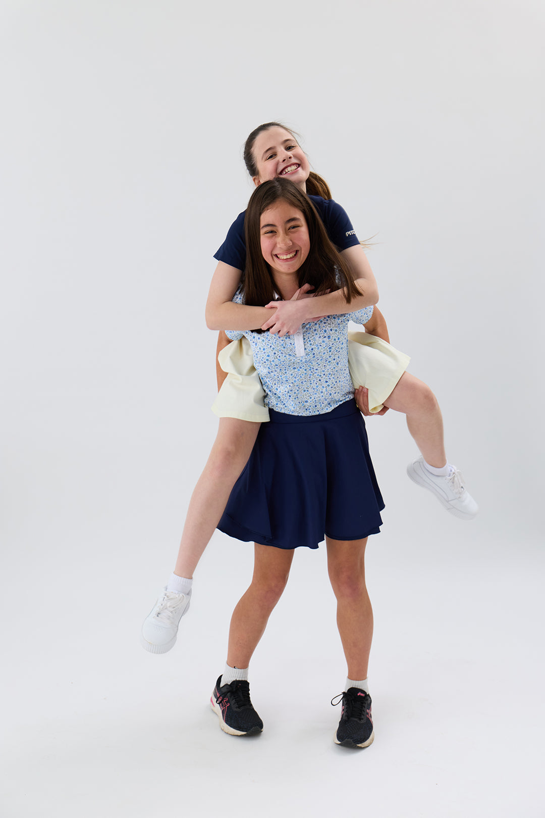 Two girls in matching navy blue and light blue golf apparel posing for a playful piggyback photo against a seamless white studio background.