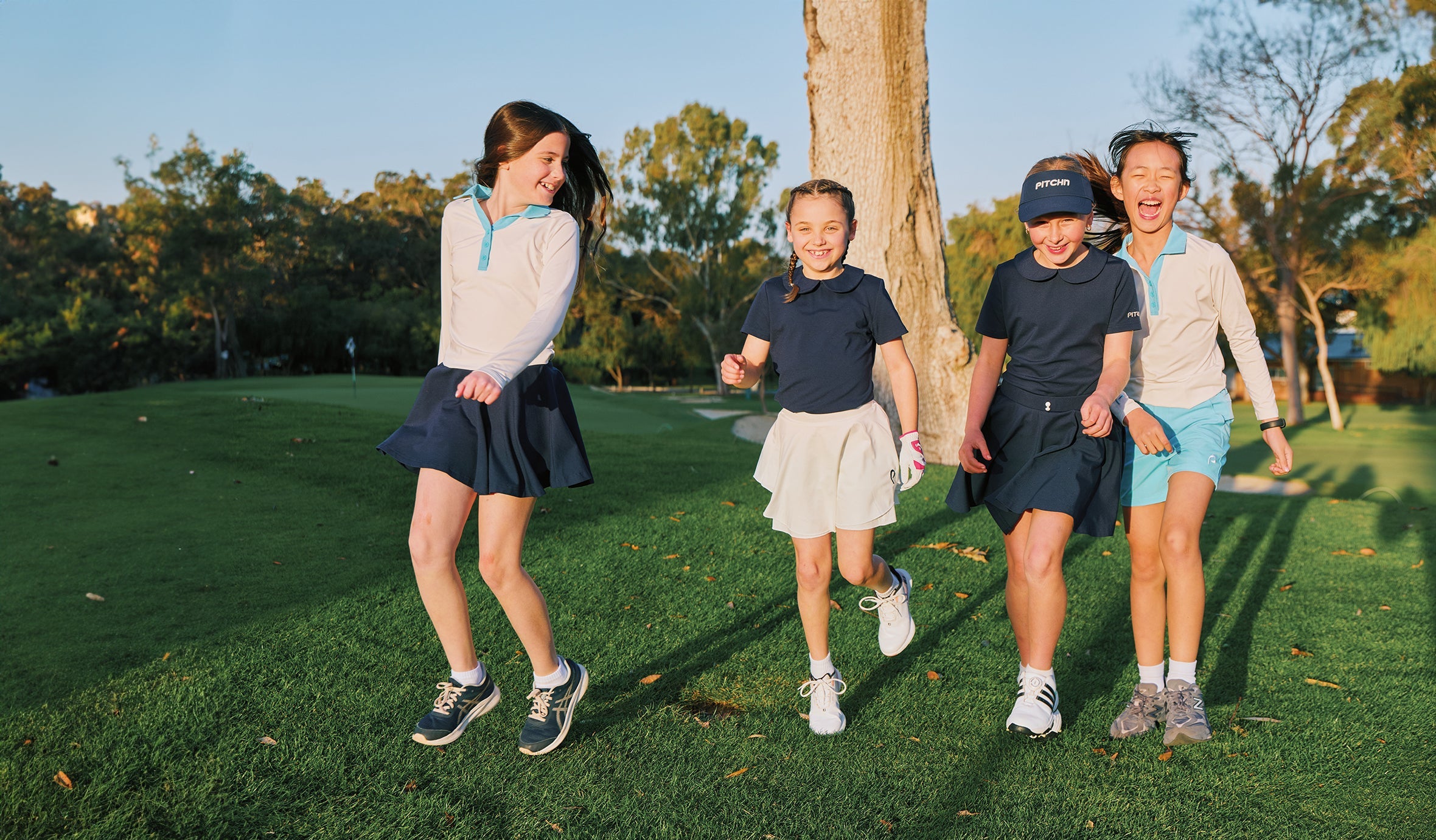 Four young girls in premium kids golf clothes are running and laughing on a sunny grassy golf course. Two girls wear navy blue and white outfits, and two wear beige and light blue outfits.